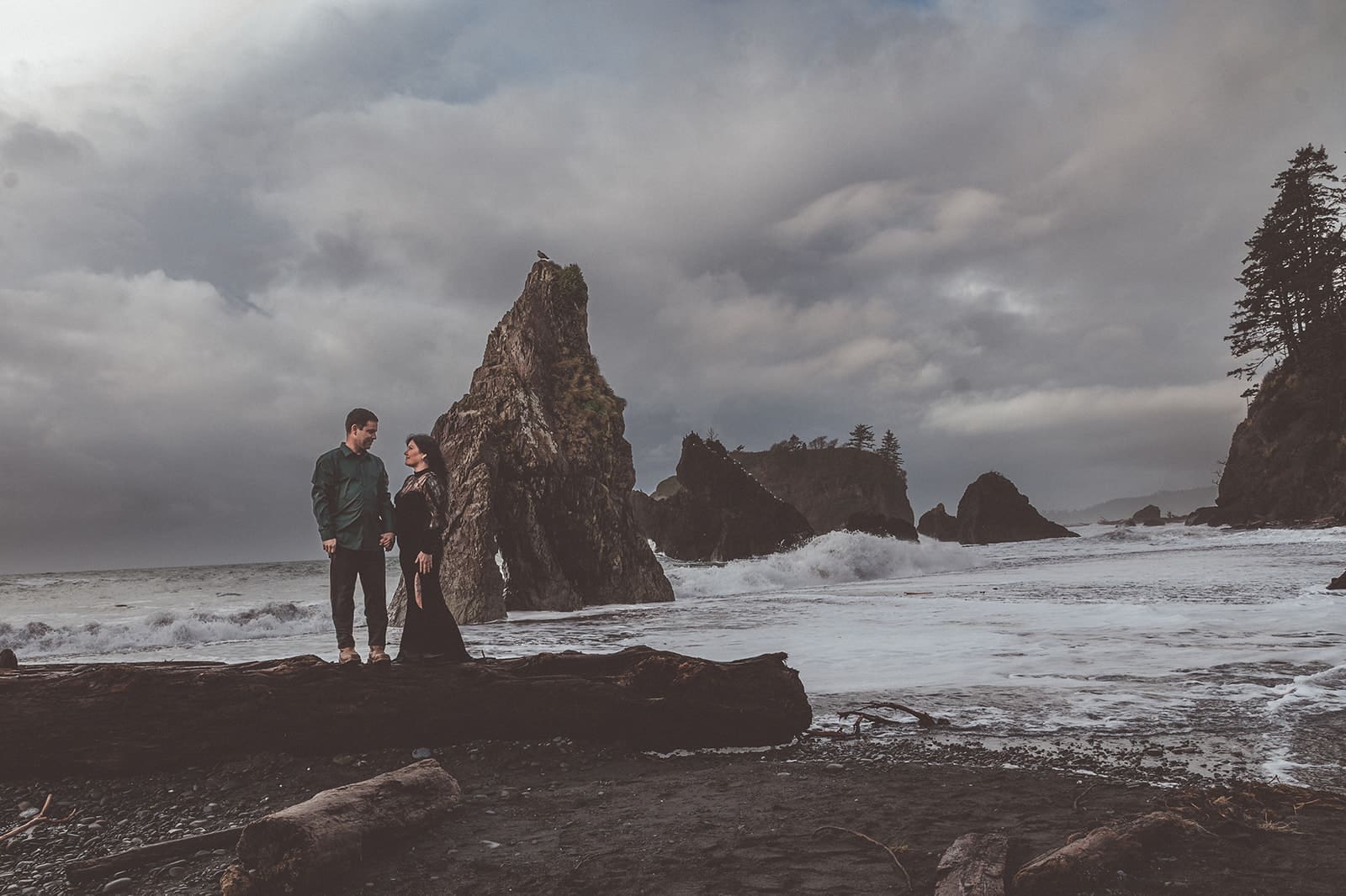couple standing on a log at Ruby Beach while looking at each other with waves and sea stacks behind them